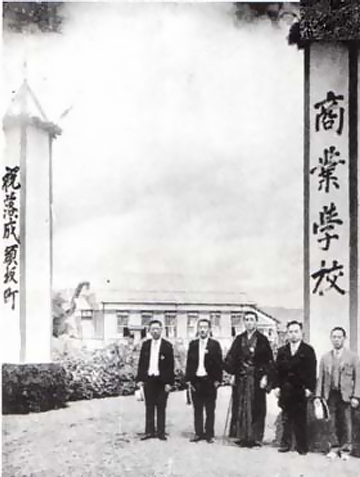 須坂商業学校落成式の写真。学校の前に、商業学校、祝落成、須坂町と書かれた大きな柱。その前に男性が5人並んでいる。