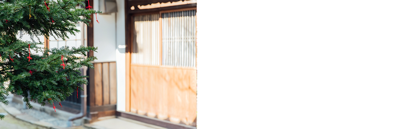 旧家の玄関か庭と思われる場所、常緑樹の枝に細くて赤い紐がたくさん結び付けられている写真。何かのおまじないだろうか?