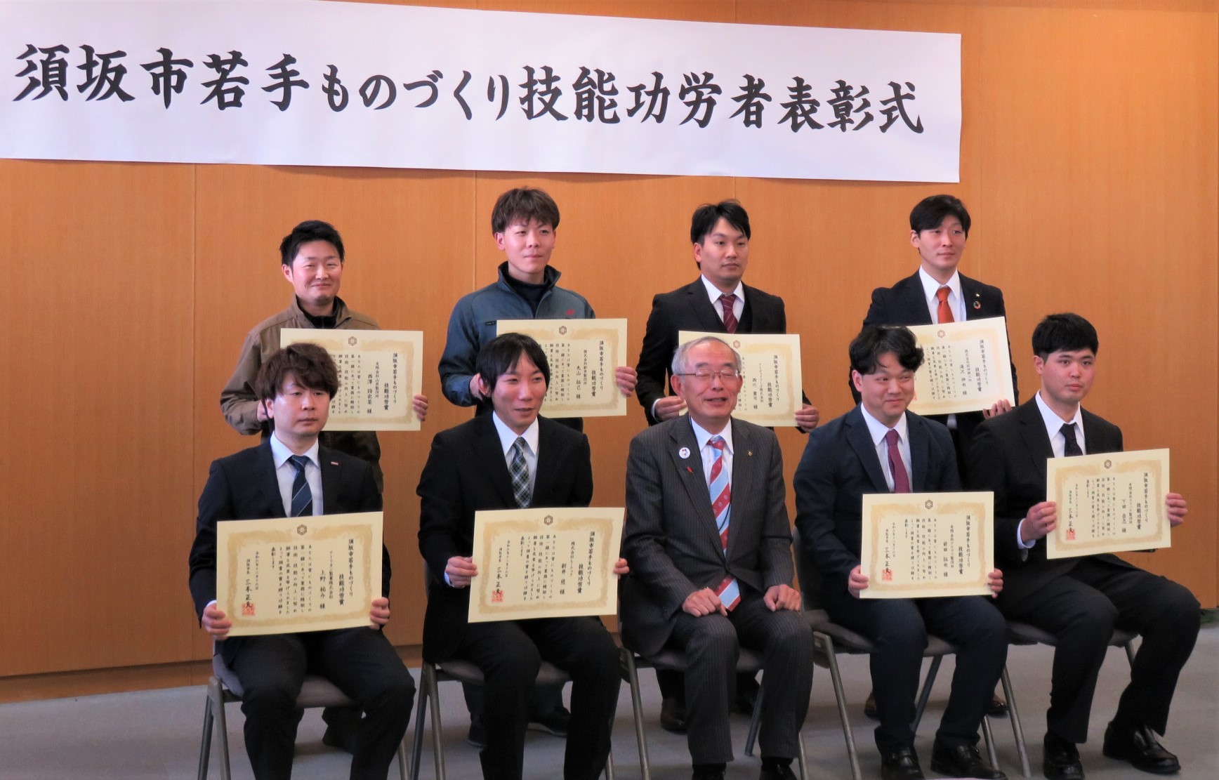表彰状を胸の高さに持った8人の須坂市若手ものづくり技能功労者と須坂市長が、横断幕の前に並んで記念撮影している様子