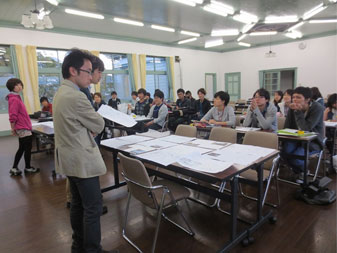 向かい合わせに2つ設置した長机の上にたくさんの資料を並べ、傍に立って見比べている先生と、席に座って様子を見ている生徒達の写真