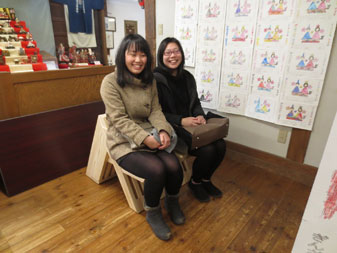 7段のひな人形が飾られた室内で、製作したベンチに女性の生徒2人が座っている写真
