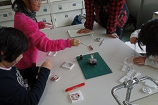 白い机の上で4名の子どもたちがアルコールランプを使用してアルミ鋳造作品を作成している写真