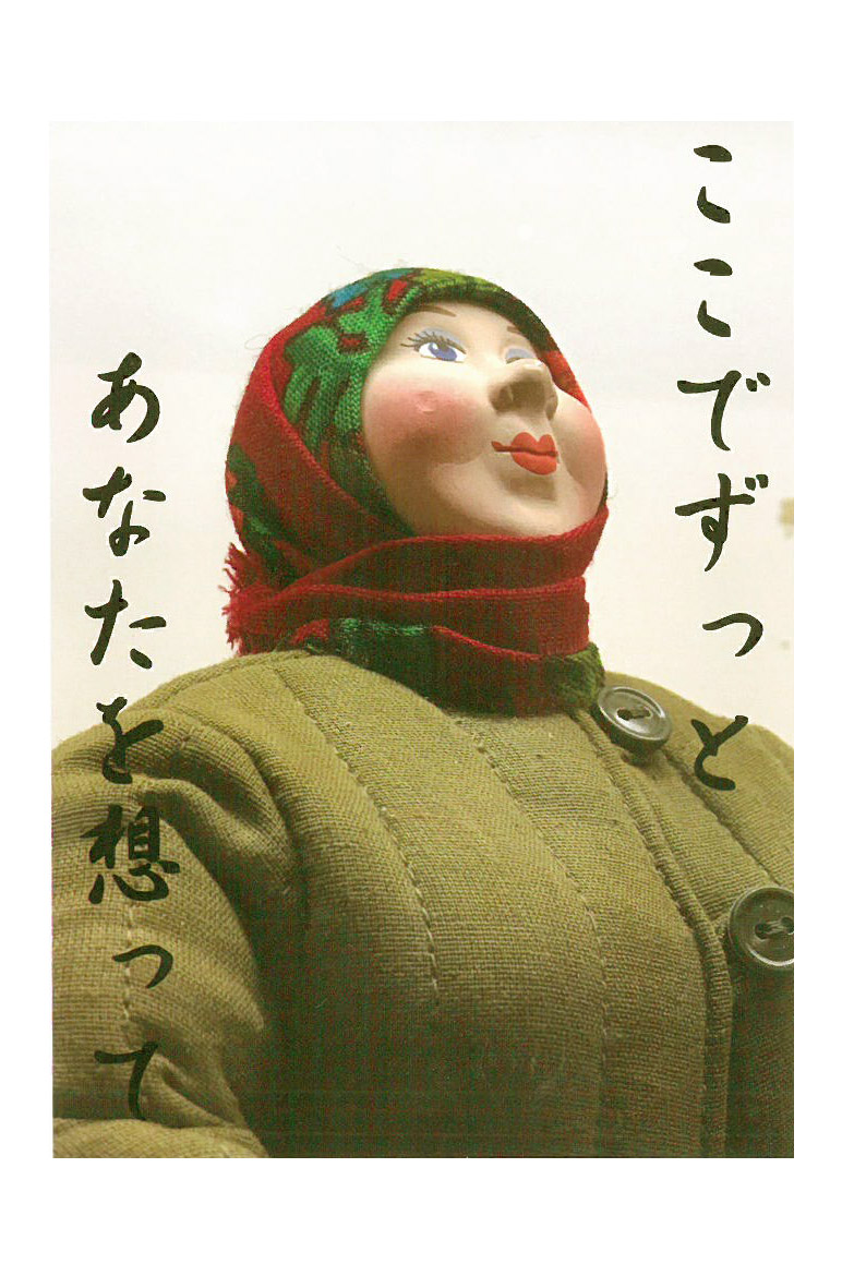ここでずっと あなたを想ってと書かれ、深緑色の服を着て、赤いマフラーを頭から巻いたふくよかな女性の人形の写真のポストカード