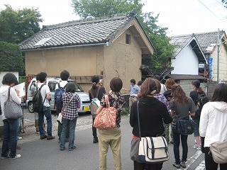 民家の外壁を示して話す男性とその周りで話を聴く大学生の写真