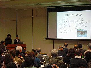 参加者がスクリーンを見ながら調査報告を受けている様子の成果発表会の写真