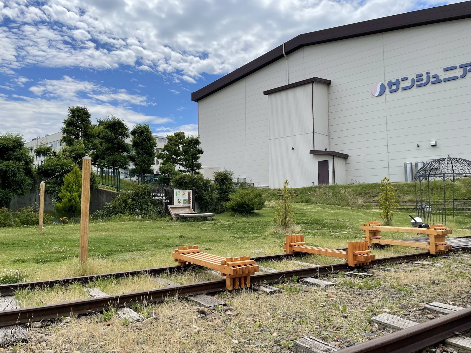建物横の芝生の広がる場所で、木を組み合わせて作られたファニチャ作品が線路上に設置されている写真
