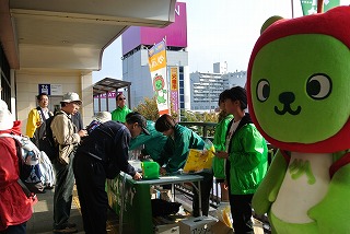 須坂駅でハイキング当日のお出迎えをする緑のジャンパーを着た学生と長野県PRキャラクターアルクマの写真