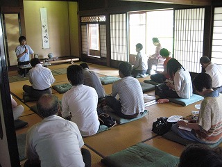 和室でマイクを持って話をしている男性と座布団に座り話を聴く学生たちの写真
