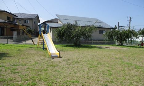 奥に複数民家があり、フェンスで囲まれた中に滑り台1台と木が2本はえている北山道公園の写真