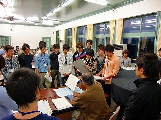 室内で長机に座った男性が話をし、男性を囲むように立って話を聞く建築学科3年生たちの写真