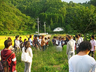 緑豊かな山付近の田んぼ道で男性の話を聞く建築学科2年生たちの写真