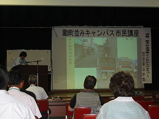 ステージ上でスクリーンに映し出された資料を見ながら話をしている男性と椅子に座り話を聴く蔵の町並みキャンパス市民講座の参加者の写真