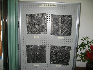 「信州大学教育学部」と書かれた紙が貼られた展示用パネルに、繭玉原型を用いたアルミ鋳造レリーフが4枚展示されている写真