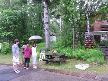 ピクニックテーブルの横で傘をさして会話する学生3名の写真