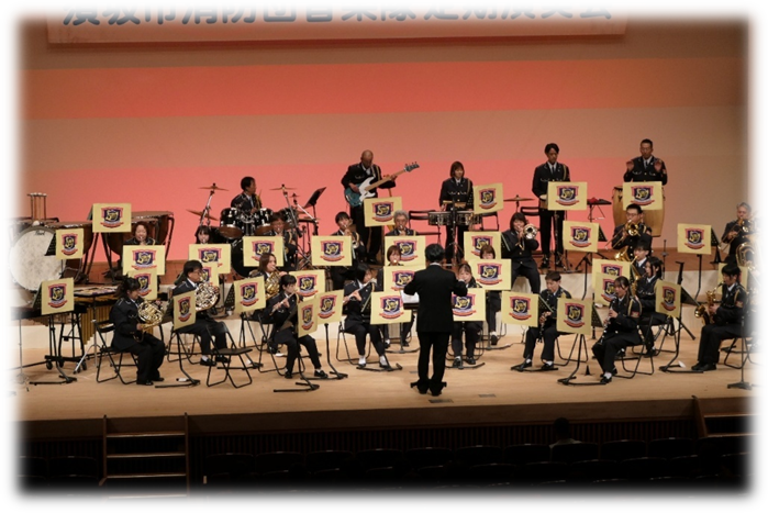 須坂市消防団定期演奏会の写真。メセナホールのステージで、指揮者を中心に、チューバ、フルート、クラリネットなど、25人ほどが演奏している。