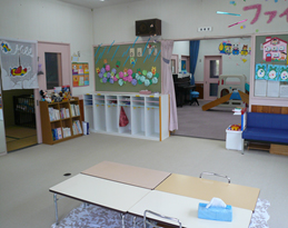 手前の部屋には4つ合わせた机が置かれ、壁にはたくさんの本が並んでいる2段の本棚やあじさいで飾られた壁面、奥には室内用の滑り台などの遊具が置かれている須坂市親子通園施設園内の写真