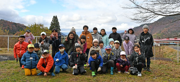 青空と山々を背景に、ダウンや帽子を被った多くの子供たちが3列に並んで記念撮影をしている写真