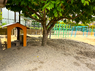 須坂保育園 園庭の一角にある手作りの木製の小屋と砂場。小屋は園児2人くらいが入れる。その横には砂場。大きな木の下にあり、子どもたちが日陰で遊べる場所
