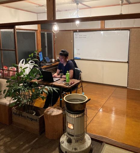 手前にリンゴ箱に入れた観葉植物とパソコン作業をする野澤さんの写真