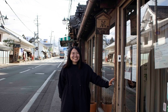 ゲストハウス蔵木製看板の前で満面の笑みを浮かべる堀口直美さんの写真