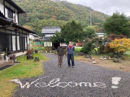古民家と庭の間にWelcomeと白く書かれた道に立つ右から若林敦さん、真由美さんの写真