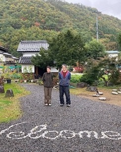 古民家と庭の間にWelcomeと白く書かれた道に立つ右から若林敦さん、真由美さんの写真
