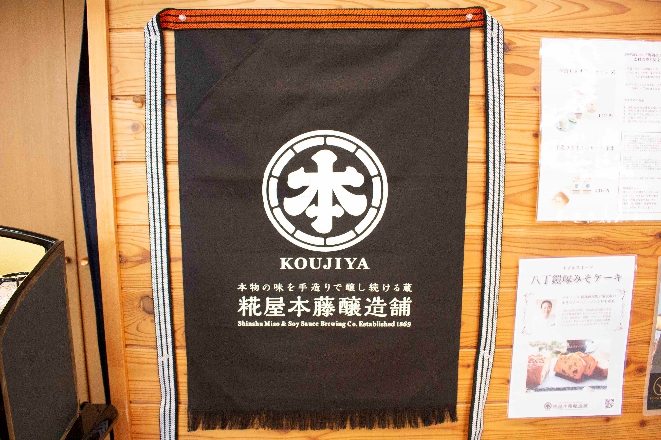 壁に貼られた白文字で糀屋本藤醸造舗と書かれた黒色の前掛けの写真