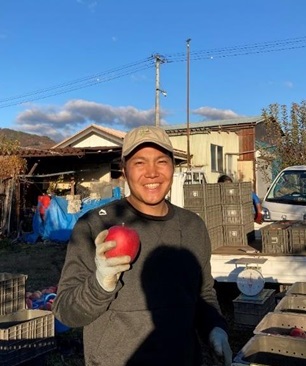 積まれたリンゴコンテナの前で撮られた右手にりんごを持ってほほ笑む藤沢拓也さんの写真