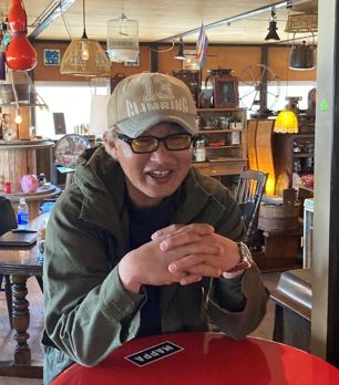 ヴィレッジ・ナニココ内で赤いテーブルに手を組む祝井一幸さんの写真