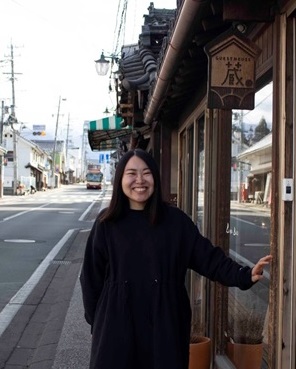 ゲストハウス蔵の木製看板の前で満面の笑みを浮かべる堀口直美さんの写真