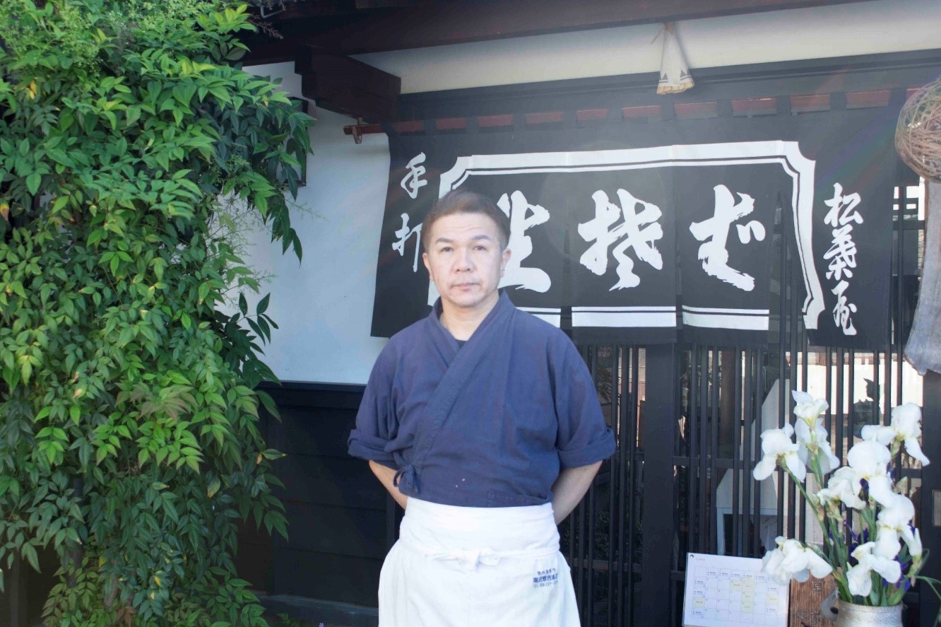 松葉屋そば店の前で両手を後ろに組み立っている松村 憲道さんを撮影した写真