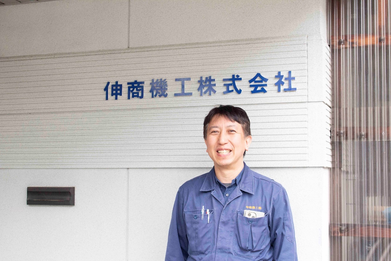 伸商機工株式会社前で撮られた紺色の作業服を着た宮川岳洋さんの写真