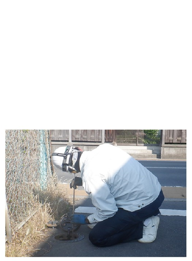 ヘルメットをかぶった職員が、道路にしゃがみ込み、丸い蓋を開けたところに長さ50センチメートル程の棒をあてて音を聴いている