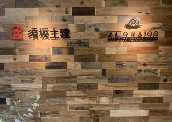 ウッドウォールパネル調の壁に須坂土建とALOHA100のロゴが貼られた写真