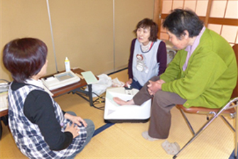 年配の女性が椅子に座って右足のかかとを機械の上にのせ、2人のエプロンを付けた女性が側で対応している健康相談の様子の写真