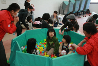 沢山のカラーボールが入った緑色のビニールプールの中で子ども3人が遊んでおり、赤い揃いのジャンパーを着た関係者の女性2名が声をかけ見守っている子育て広場の様子の写真