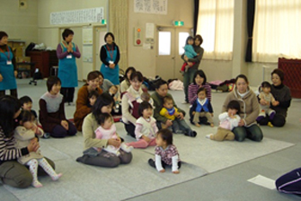 小さな子どもを膝の上に抱いた親子連れの皆さんが沢山座っており、青いエプロンを付けた関係者の方々が後ろに立って見守っている子育て支援の様子の写真