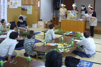 前方の檀上でお面を頭に着けて紙芝居を披露している4名の女性と、設置された複数のテーブルに参加者たちがが座って紙芝居を楽しんでいる様子の写真