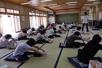 広々とした和室に参加者の皆さんが間隔を開けて座り、片足を前に出して両手を出した片足のつま先まで伸ばしてストレッチをしている写真