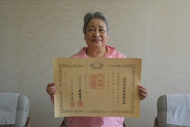 保健補導員会の関係者の女性が春の緑綬褒章の賞状を広げて持ち、写っている記念写真