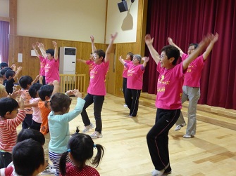 ピンクのお揃いのTシャツを着た保健補導員8名が、参加している子供たちの前に立って、足踏みをしながら両手を上に上げ、子供たちが保健補導員と同じ動きをしている写真
