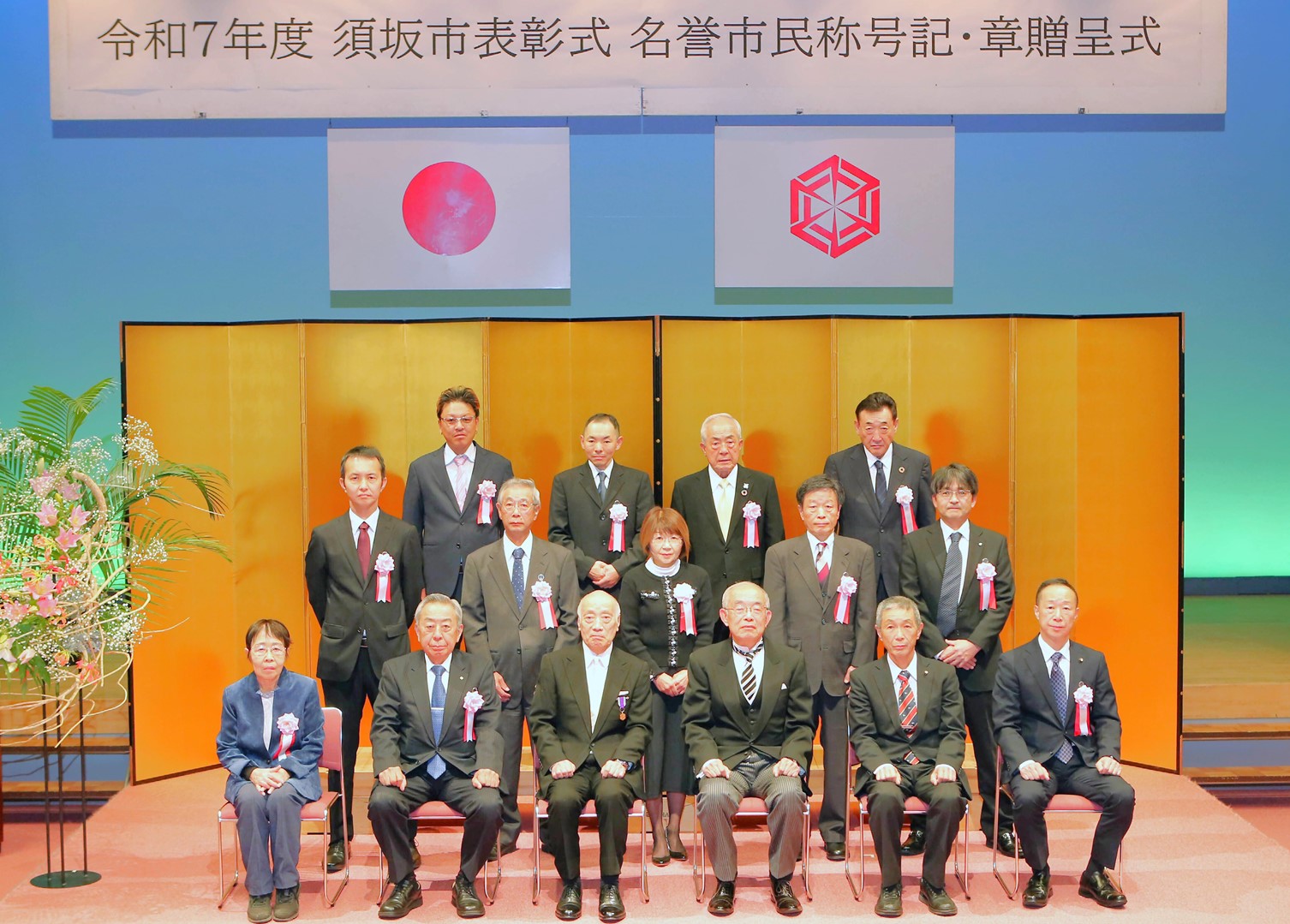 「須坂市表彰式、名誉市民称号記・章贈呈式」と書かれた横断幕、国旗、市章旗が掲げられた空間で、金屏風の前で表彰受賞者と市長が写る集合写真