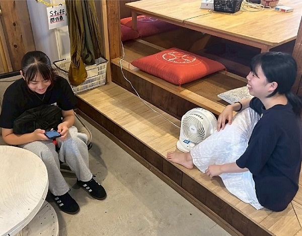 女子高校生が椅子に座り、手に持ったスマートフォンを見ながら笑顔を浮かべている。その向かいに女性が座り、女子高校生に向かって笑顔を浮かべている。