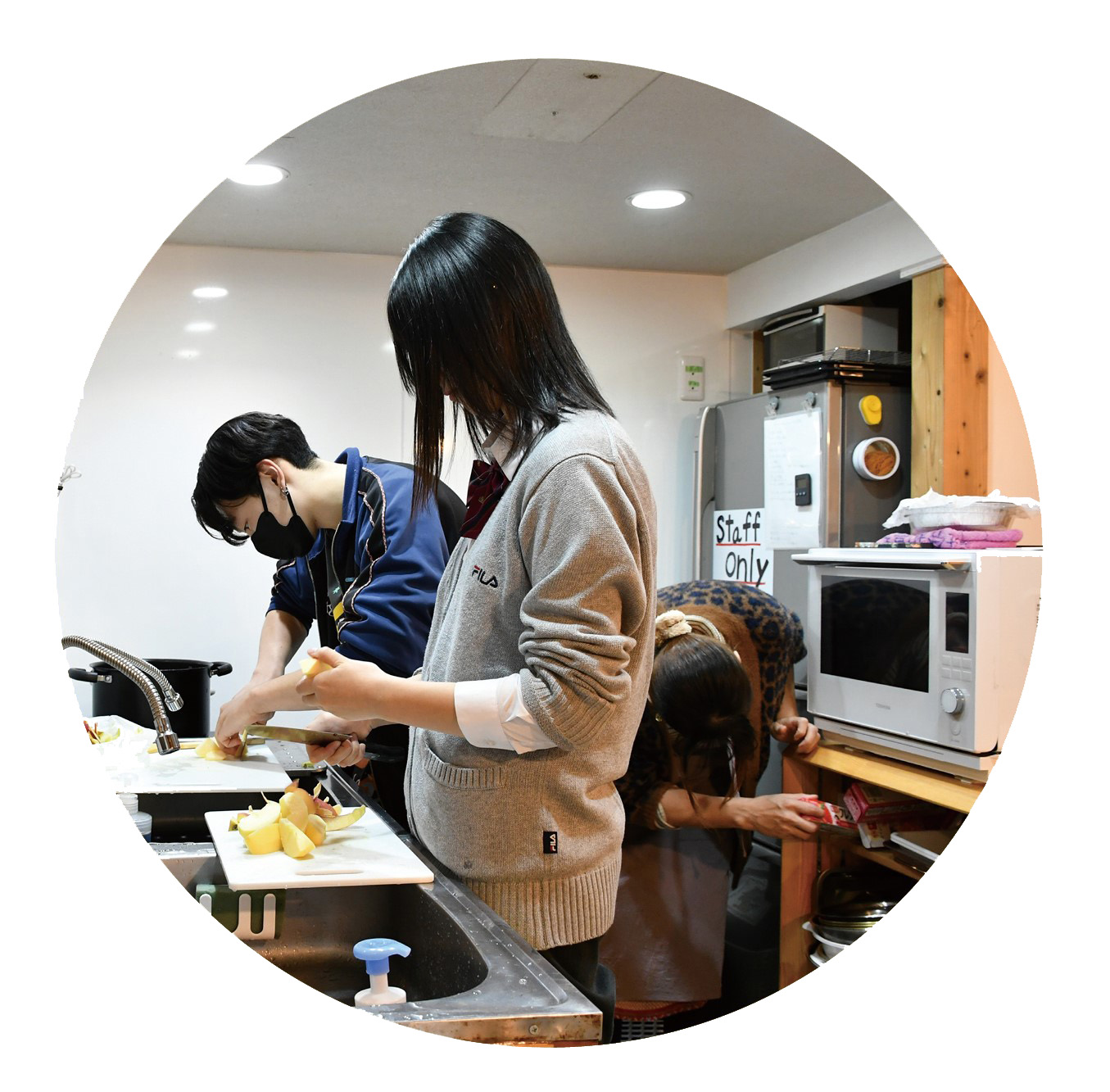 シンクに向かい、まな板でリンゴを切っている男子高校生とリンゴを向いている制服姿の女子高校生。冷蔵庫、オーブンも見える。