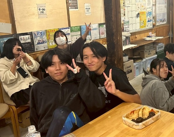 男子高校生2人と女子高校生1人、女子大学生のユースワーカー1人がこちらに向かって笑顔でピースサインを向けている。制服を着た1人の女子高校生は口にお菓子を入れようとしており、机の上にはお菓子が置かれている。