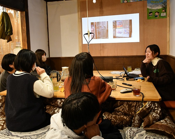 大きなこたつを囲んで、女性と高校生5人が話をしている。奥の壁には画像が投影されている。