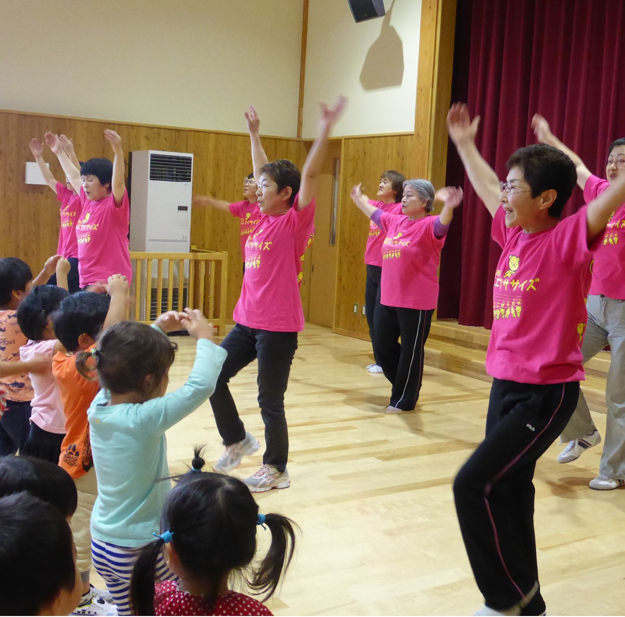 揃いのピンク色のTシャツを着た男女8人が、室内で園児に向かって、両手を広げて上に上げ、足踏みをするようなポーズの体操を披露している。園児も真似をして両手を上げている。