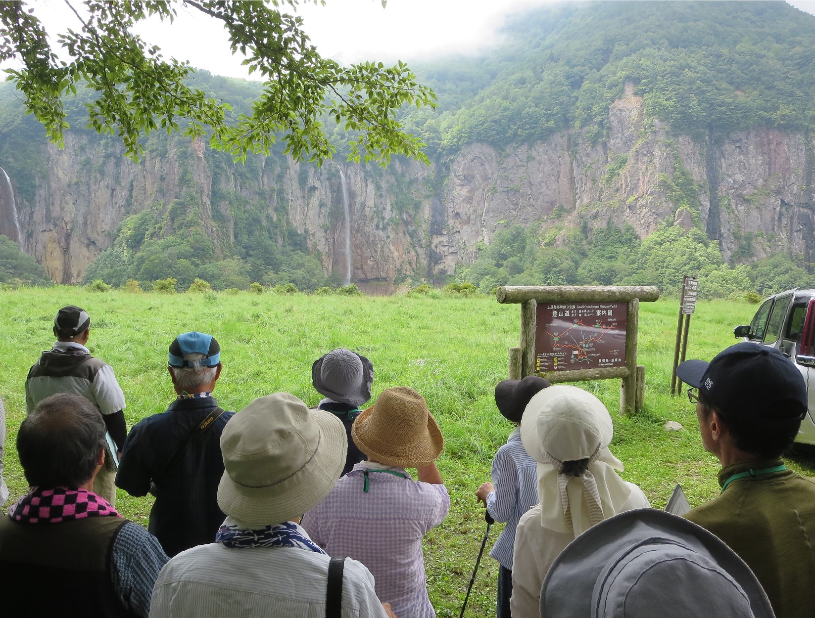 米子瀑布群が正面に見え、手前に鉱山跡地の名残を示す平地が広がる場所に、参加者男女10人の後ろ姿が写る。