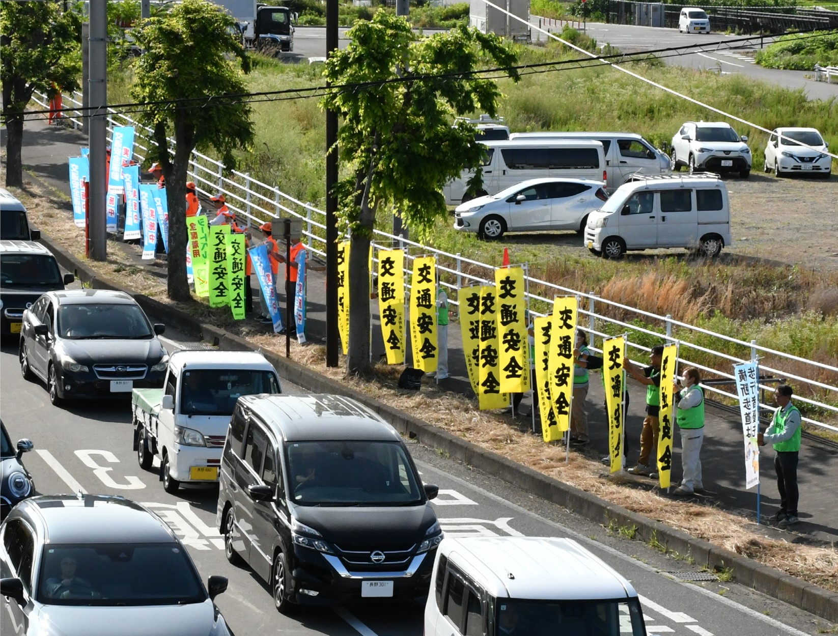 車道で多くの車が信号待ちをしている横で、20人位の人が歩道に並び「交通安全」と書かれたのぼり旗を持って交通安全の呼びかけをしている。