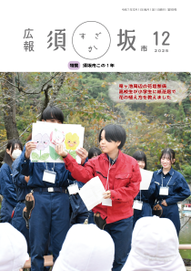 広報須坂12月号の表紙写真「竜ヶ池周辺の花壇整備、高校生が小学生に紙芝居で花の植え方を教えました」屋外で作業着を着た高校生たちが花の絵が描かれた紙芝居を演じている。手前に白い帽子をかぶった小学生の後ろ姿が見える。